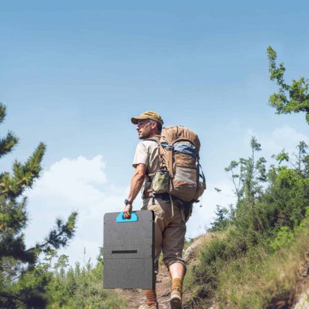 Backpacker hiking on a mountain trail with a large backpack, carrying a portable speaker, amidst lush greenery and clear blue sky, embodying off-grid adventures and outdoor exploration.